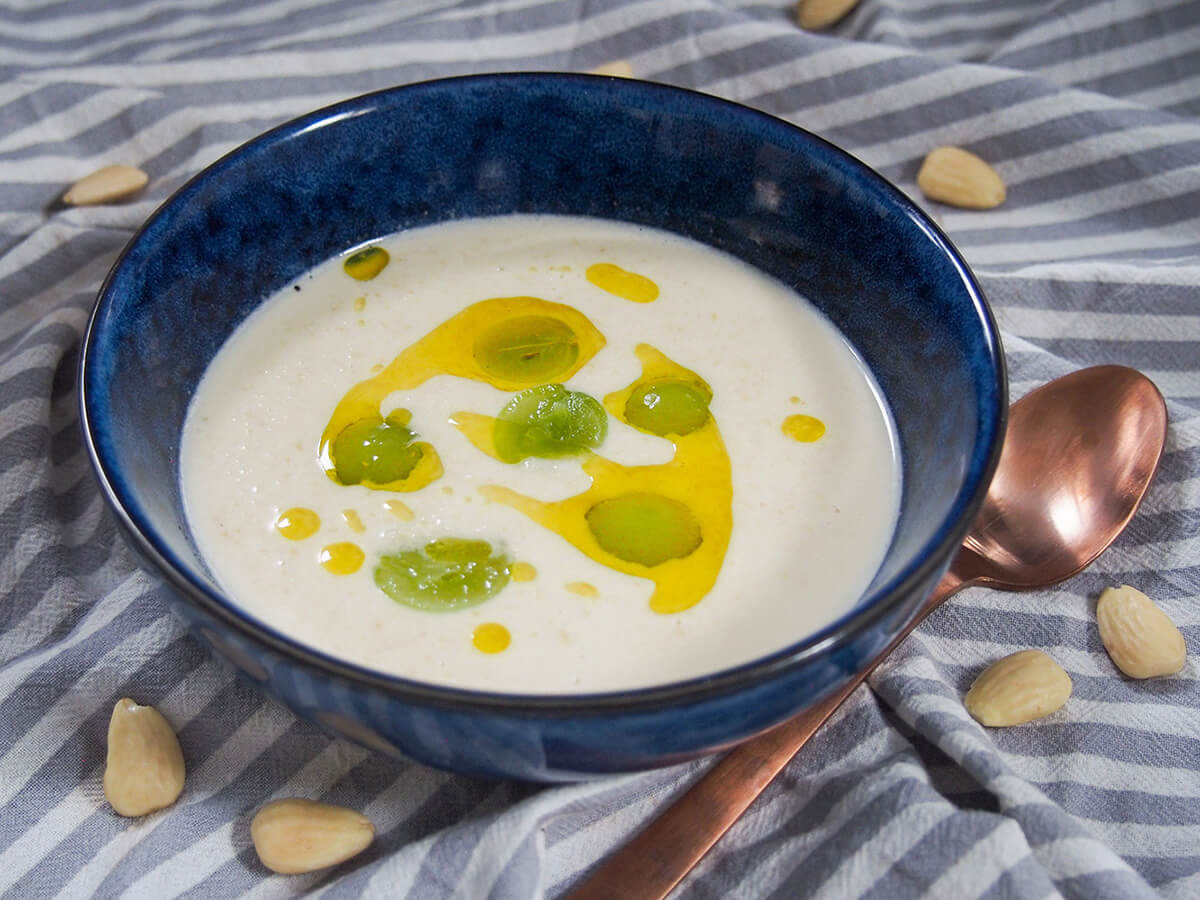 Ajo blanco is a wonderfully light cold Spanish soup made with almonds and bread. It easy to make, has delicate flavors and is gently filling yet refreshing. Perfect for a warm summer's day. bowl of ajo blanco with spoon to side and grapes garnishing soup