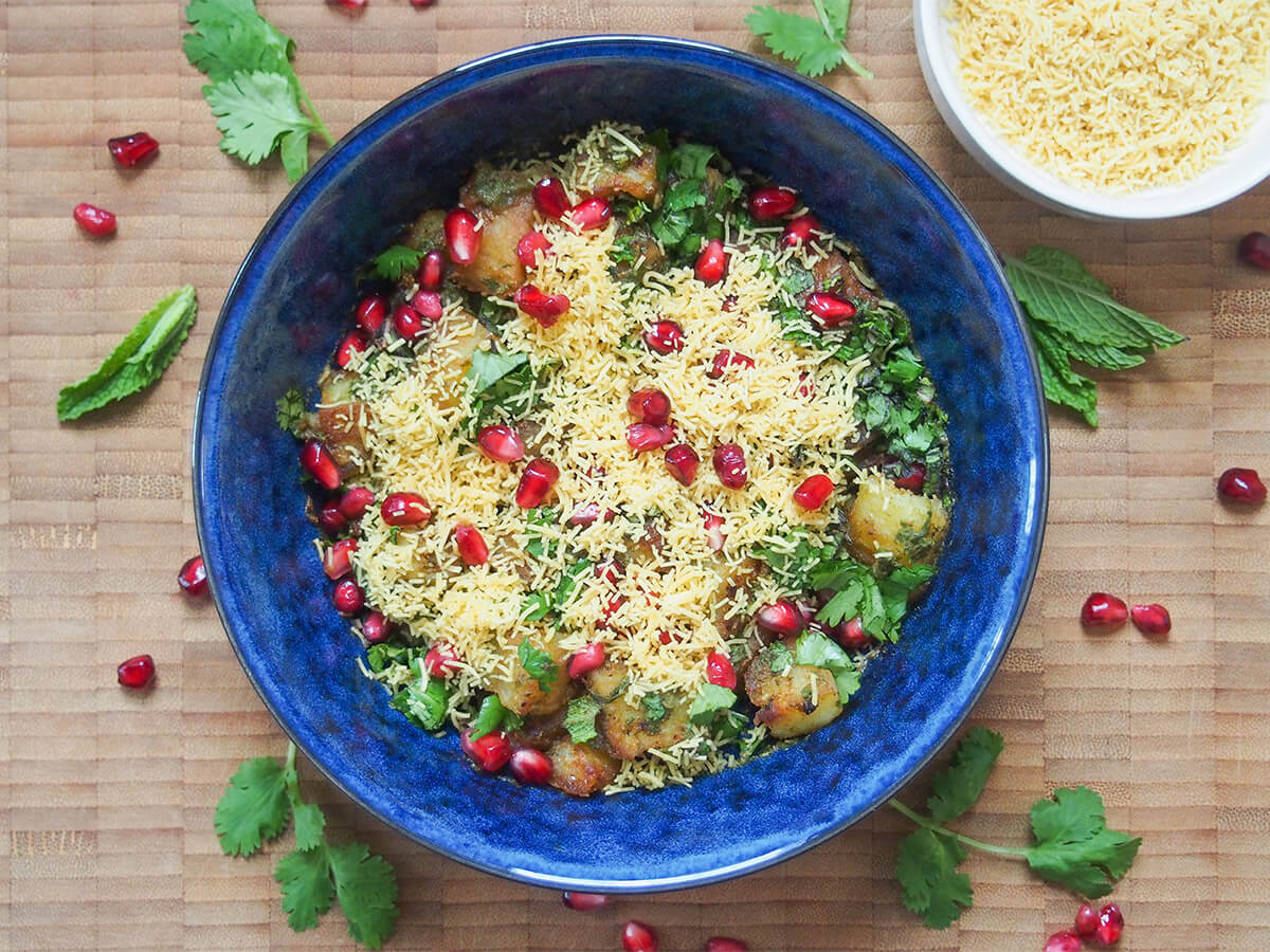 Aloo chaat is a delicious Indian snack combining fried potatoes with spices, chutney, herbs and more. It's easy to make, has tangy and spicy flavors and is addictively good. bowl of aloo chaat with additional sev to side and herbs and pomegranate around bowl