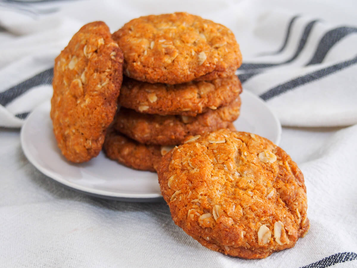 Anzac biscuits are a popular Australian sweet treat with a doze of history. These eggless biscuits/cookies are easy to make with a delicious mix of oat and coconut flavors - definitely worth enjoying soon. stack of Anzac biscuits with one in front on plate and one rested against stack