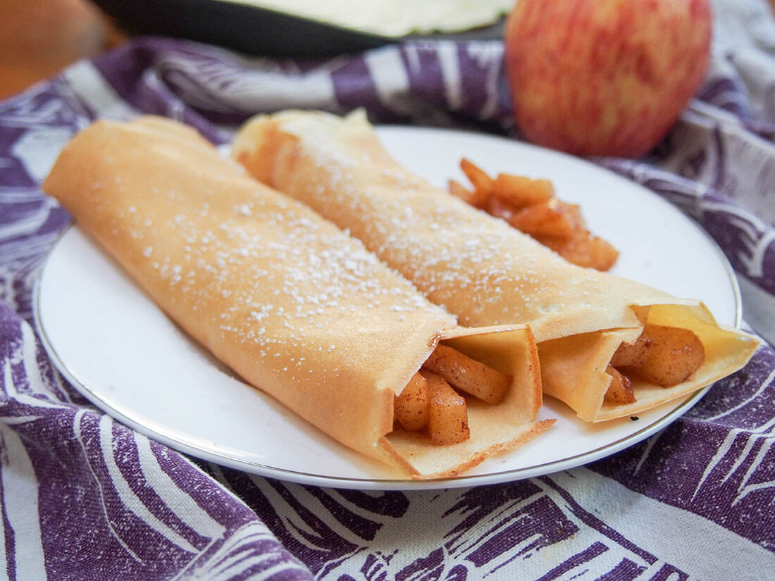 Light crepes filled with a gently sweet, cinnamony apple filling, these apple crepes are a delicious taste of fall. Easy to make, and perfect for dessert, brunch, or a tasty snack. apple crepes on plate with apple behind