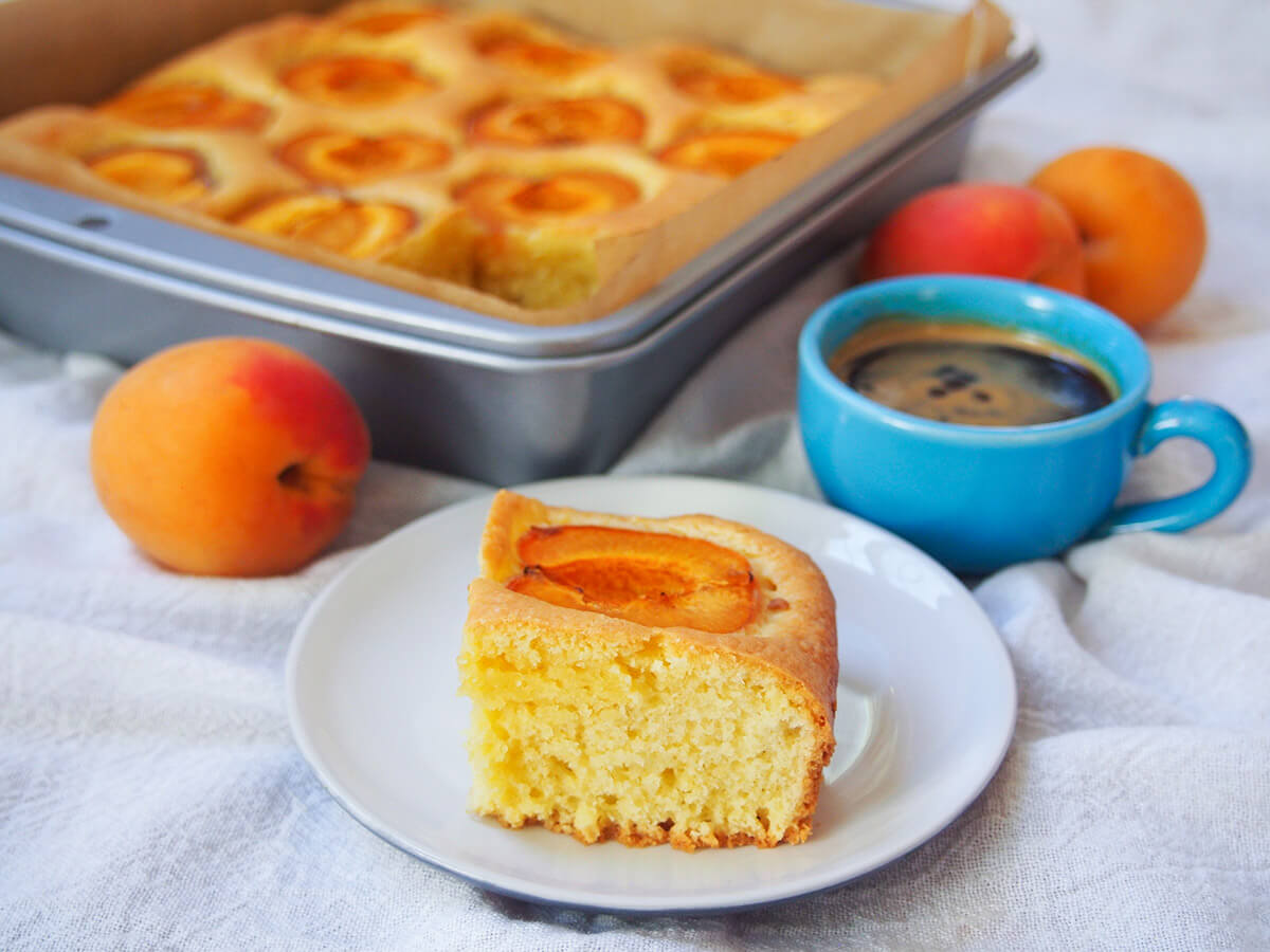 Austrian apricot cake is a wonderful combination of a simple cake base, with a touch of lemon, with large pieces of tender apricots nestled on top. It's a popular choice during the fruit's season alongside coffee, and makes a delicious anytime treat. slice of apricot cake marillenkuchen on plate in front of tin with rest of cake, coffee cup to side of tin