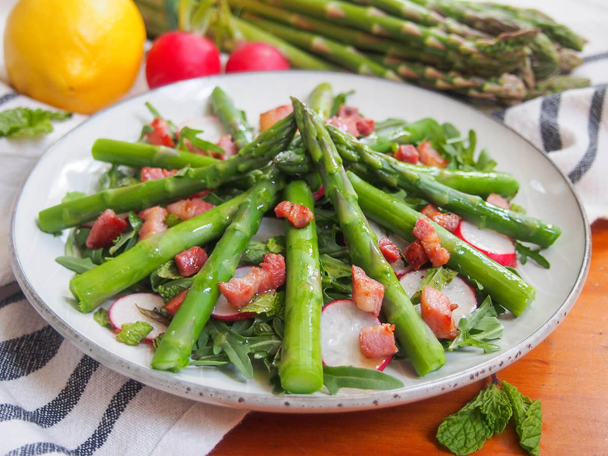 This easy asparagus salad is full of the tastes of spring, with asparagus, greens and radishes topped off with a bright lemon vinaigrette and crisp pancetta. It's perfect as a side or appetizer for a spring time meal. side view of asparagus salad with lemon vinaigrette with asparagus spears stacked into middle of plate