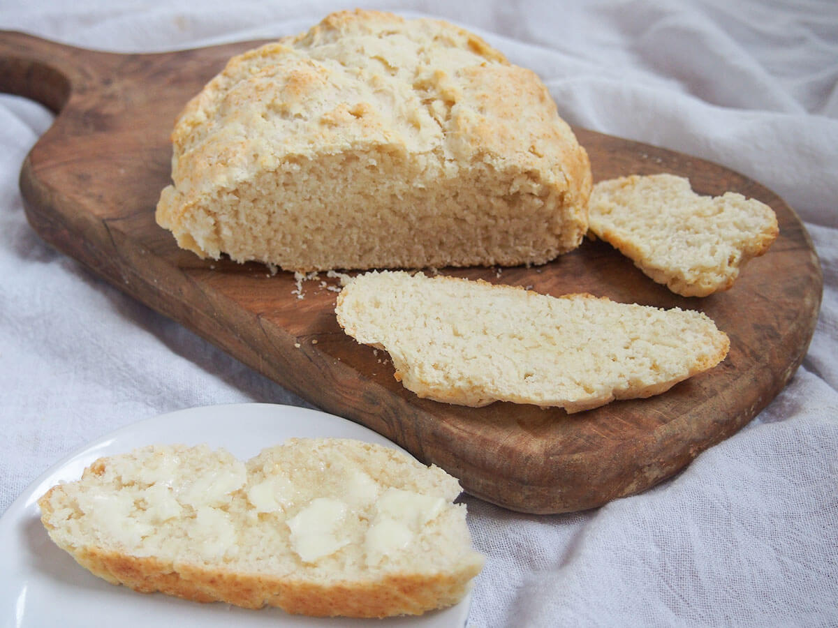 Australian damper is a type of soda bread that's really quick & easy to make. Traditionally cooked in the fire, it's just as easy at home. Great with sweet or savory toppings. slice of Australian damper bread with butter on plate in front of loaf and slices on board