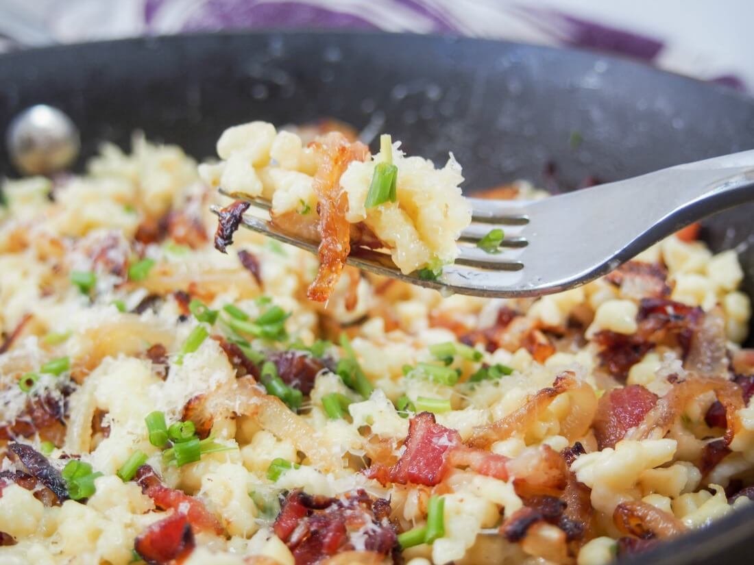 Bacon onion spaetzle in a simple but delicious combination of spaetzle (or other small pasta), bacon, caramelized onions and cheese. Perfect comfort food, it makes a delicious lunch. Taking forkful of bacon onion spaetzle from skillet