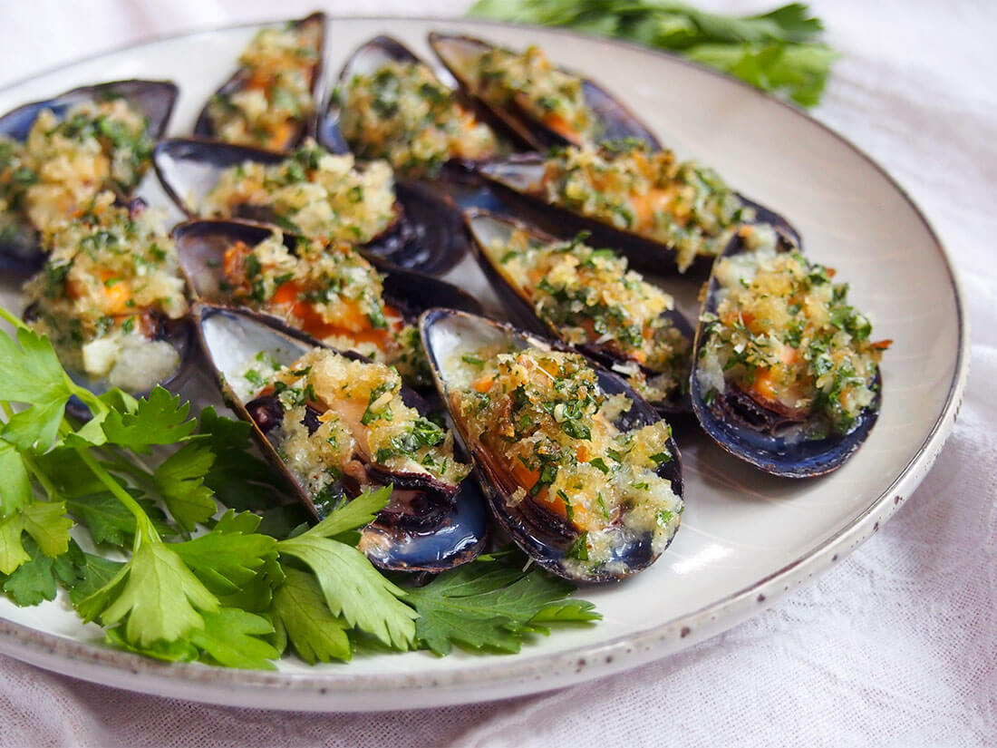 These baked mussels are easy to prepare and make a great appetizer for any occasion. Tender mussels with a tasty garlicy crumb topping - they're elegant finger food (that's budget-friendly too). baked mussels on plate with parsley garnish to side