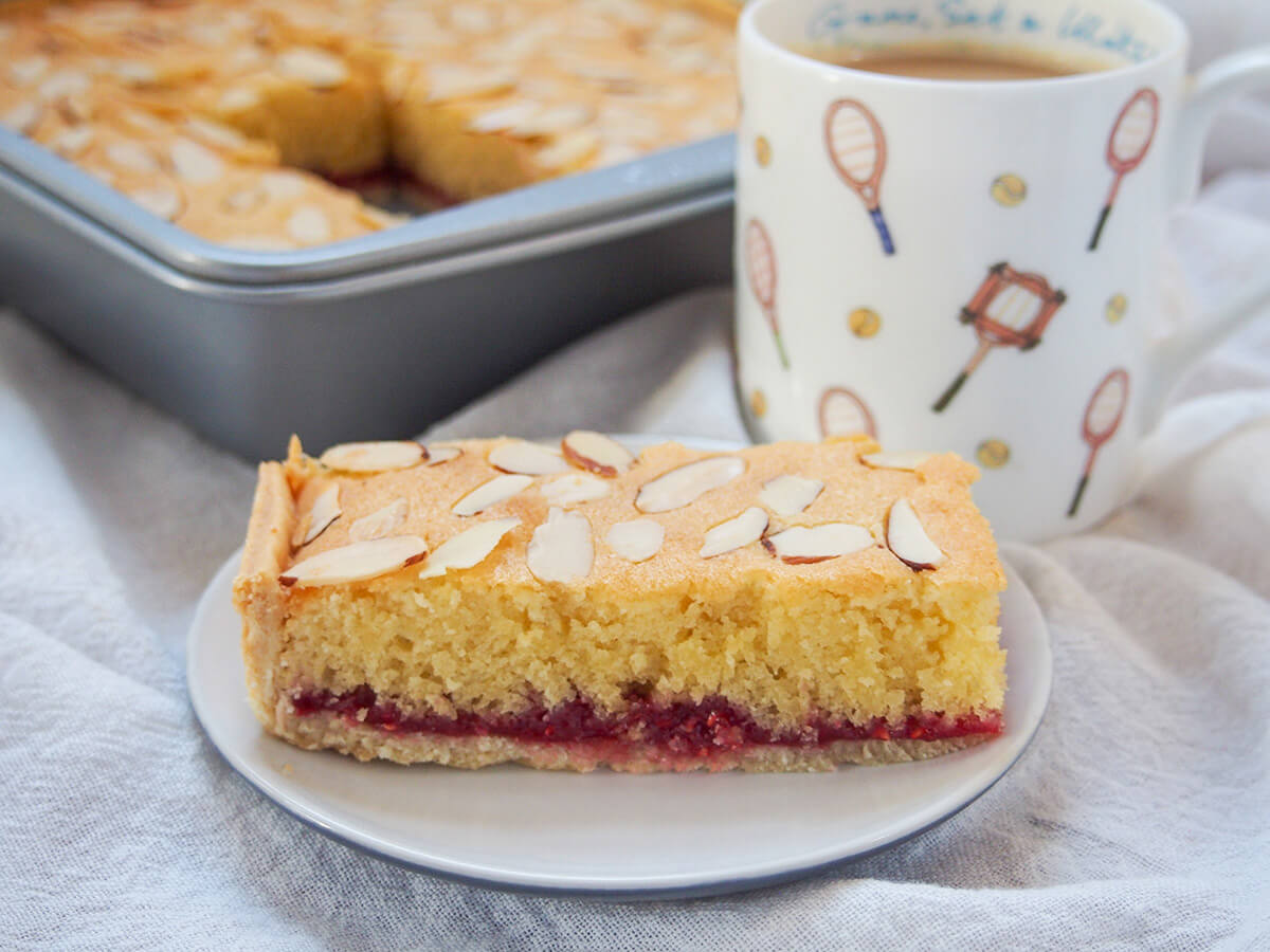 Bakewell slice is a classic British tea time treat, with layers of pastry, raspberry jam and an almond sponge-like top. It's easy to make and a tasty combination sweet and gently tart flavors. piece of Bakewell slice on plate with mug behind