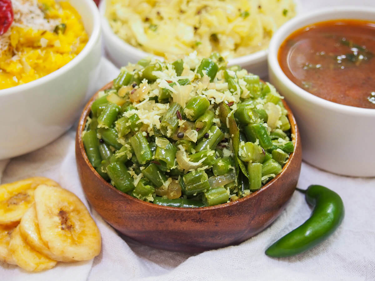 Beans thoran is a popular Keralan side dish that's quick and easy to make. These stir fried beans with coconut, green chili and simple spices are light, tasty & versatile. dish of beans thoran with chili to one side and banana chips of other with other dishes behind