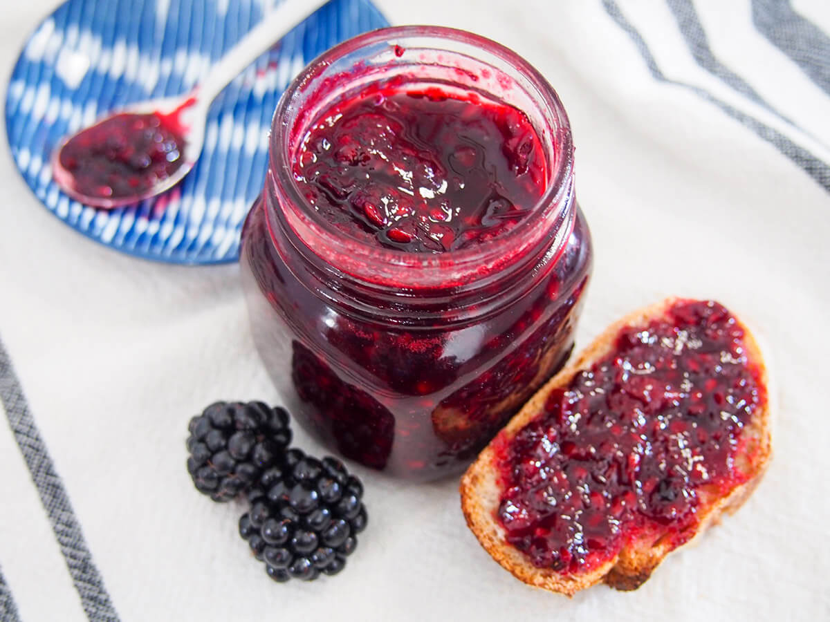 Blackberry jam is brightly colored and bursting with fruity flavor. This one is made in a small batch with lower sugar and comes together quickly for a delicious spread, perfect for toast and more. jar of blackberry jam with some on bread to one side and spoonful on small plate behind