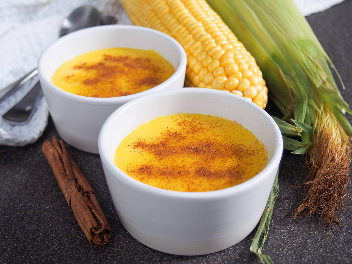 This Brazilian corn pudding, curau de milho, is a simple custard or pudding-like dessert that you can enjoy warm or chilled. It's easy to make with a bright color, smooth texture & tasty corn flavor. two dishes of Brazilian corn pudding with corn behind and cinnamon stick to side