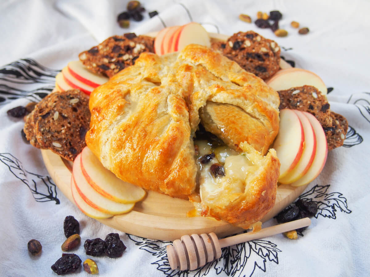 Brie en croûte is a wonderfully simple and decadently delicious appetizer or cheese board addition that is sure to please any cheese lover. It's simple to prepare, adaptable and perfectly gooey and tasty. board with brie en croute, apple slices and crackers around and dried fruit and heney dripper around board.