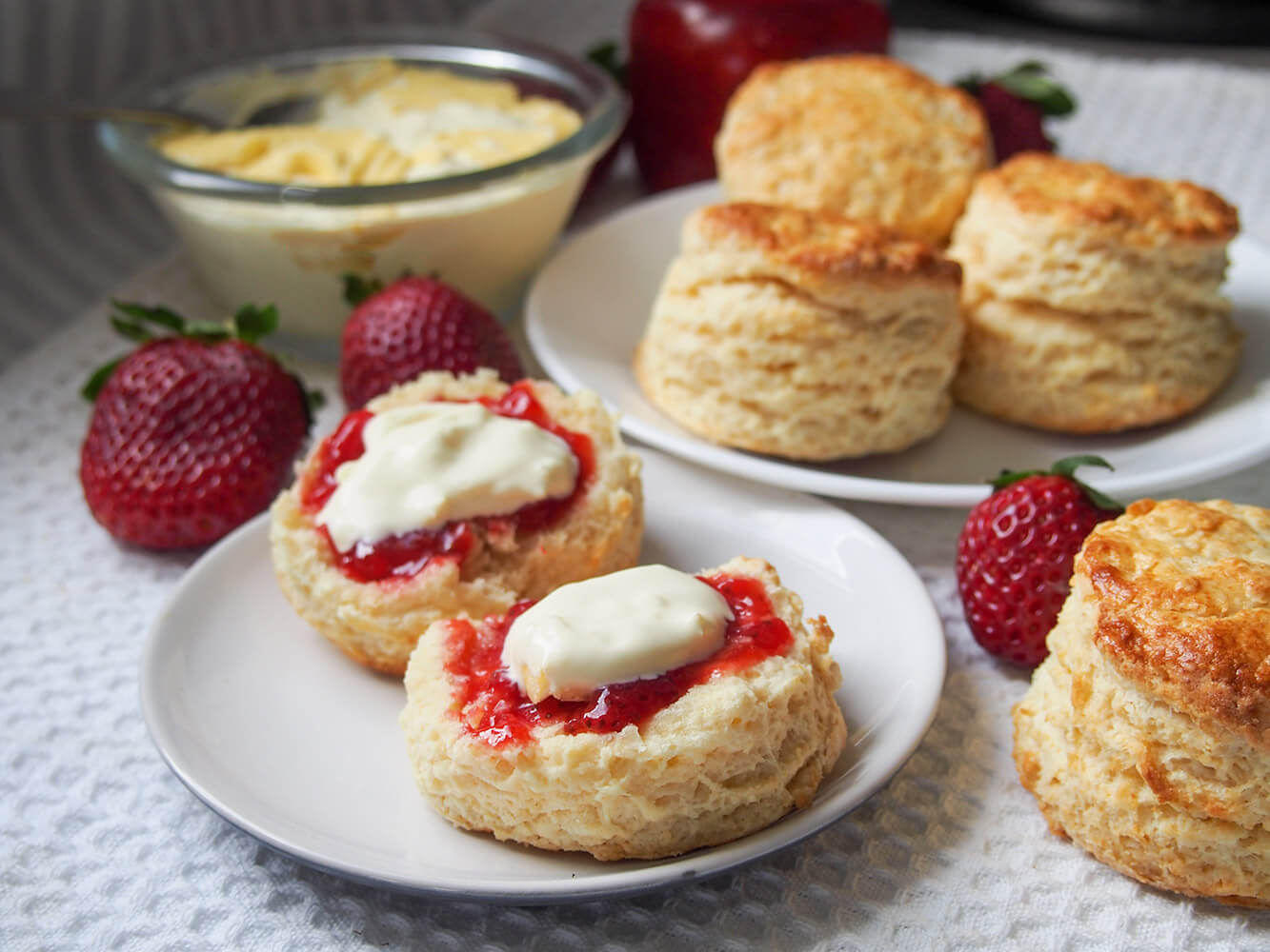 British scones are a classic treat to enjoy as part of a 'cream tea' with jam and cream. They are easy to make, wonderfully soft and gently sweet. Delicious in every bite. a scone split open and topped with jam and cream with more whole scones on plate behind