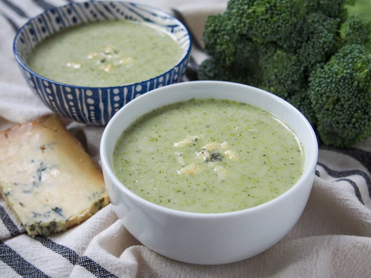 Broccoli and stilton soup is a wonderfully tasty, comforting soup that's flavorful and easy to make. Just a few ingredients & a short cooking time, this blended soup is light yet gently rich goodness. Perfect for lunch/as a starter. side view of two bowls of broccoli and stilton soup with stilton wedge to side and broccoli behind.