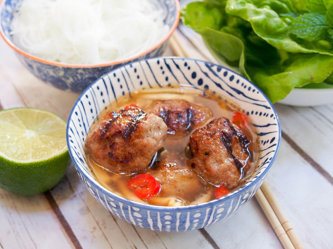 Bun cha Hanoi is a traditional Vietnamese dish that is so worth getting to know. A delicious combination of flavorful meatballs, rice noodles and a tangy dipping sauce/broth, you'll immediately love all the flavors and textures. Bun cha Hanoi bowl of meatballs with noodles and salad behind