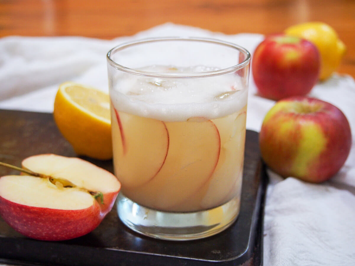 This calvados sour takes the classic, simple tart and strong formula and gives it a fresh, slightly autumnal twist. It has a lovely balance of flavors including apple and a little spice. glass of calvados sour with cut apple to side and apples and lemon behind