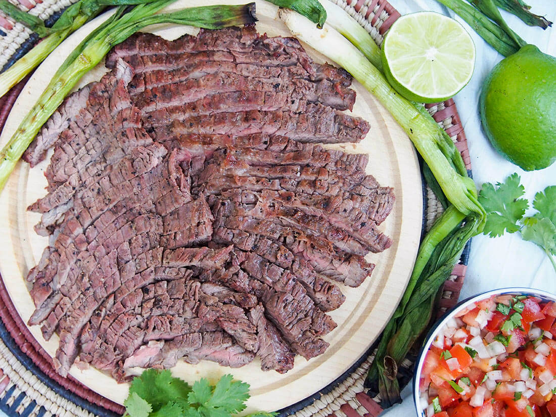 This traditional Mexican carne asada is a delicious and easy marinated, grilled beef dish that makes tacos perfect for summer. With minimal ingredients and effort, it's a meal you'll have on repeat. traditional carne asada grilled beef on board with grilled green onions and pico de gallo to side