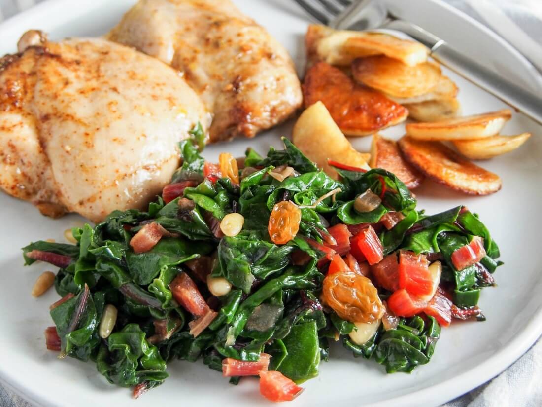 Catalan-style sauteed Swiss chard with raisins and pine nuts is an easy and delicious way to serve greens as a side dish Catalan-style sauteed Swiss chard with raisins and pine nuts on plate with chicken and pan-fried potatoes behind