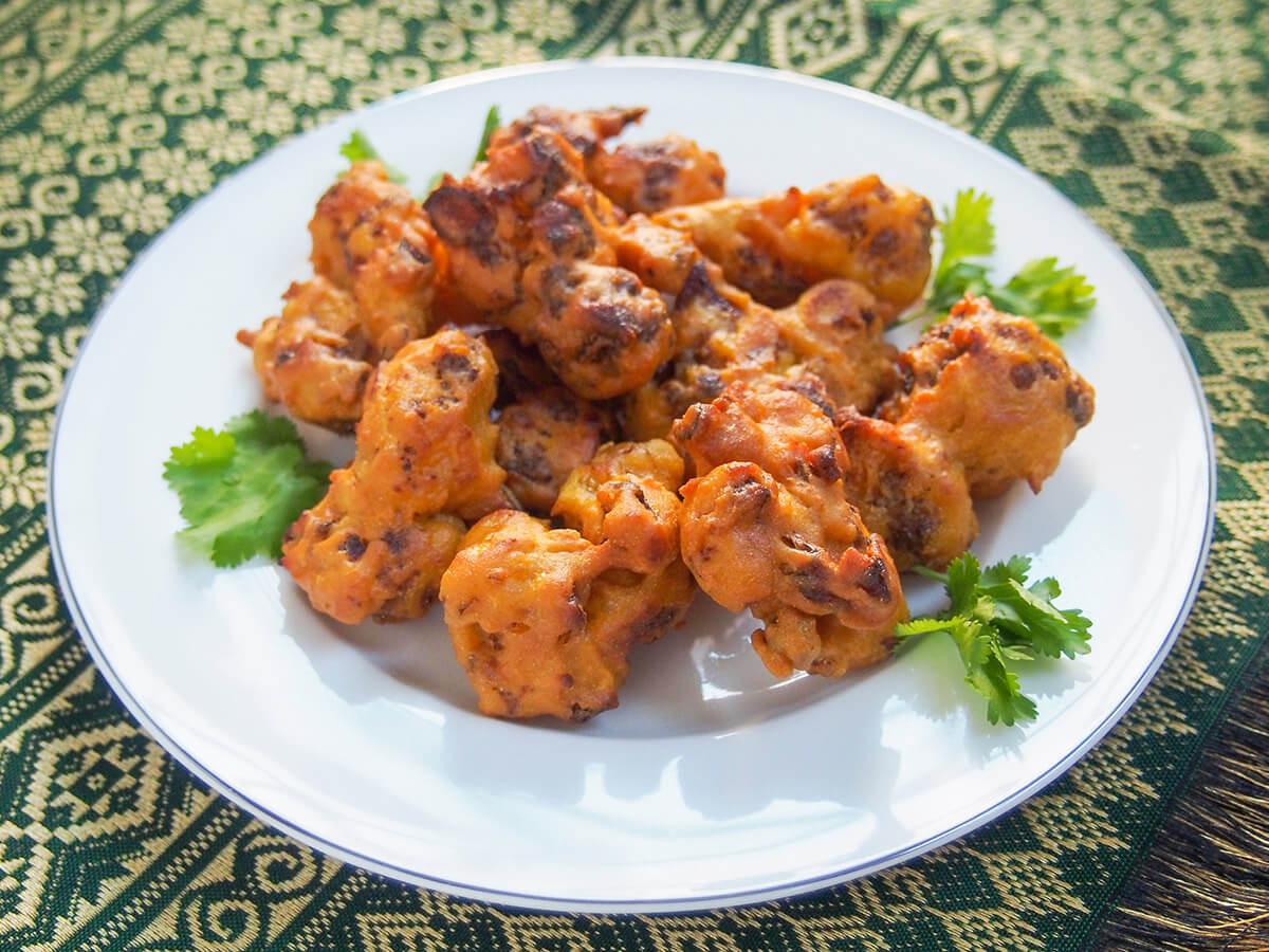 Cauliflower pakora are a traditional Indian snack or appetizer that's both tasty and versatile. Cook up some of these fritters to start your next Indian meal or add some to your game day or party menu! plate of cauliflower pakora on green and gold cloth
