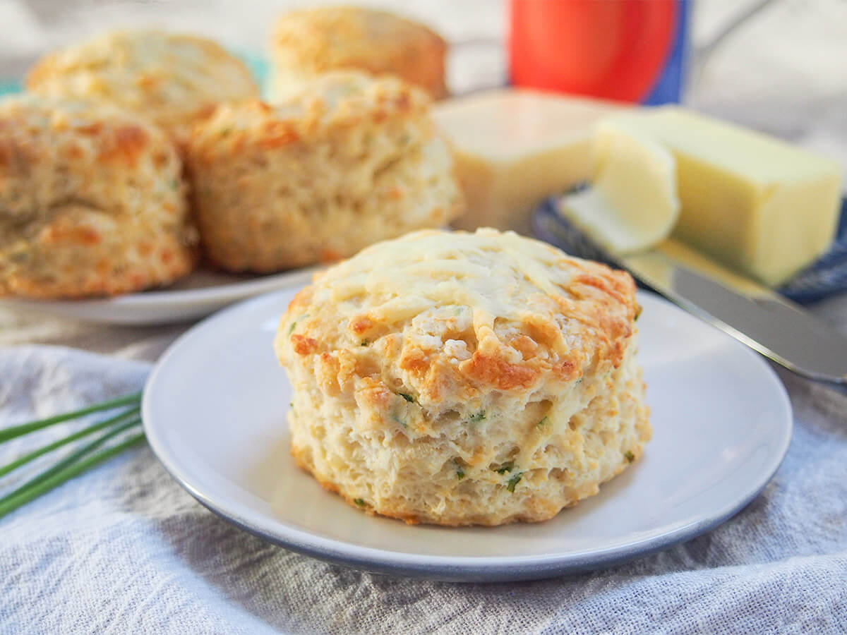 These savory cheese scones are easy to make, packed with cheese flavor yet surprisingly light. A delicious snack, side or base for a sandwich. a cheese scone on a small plate with another plate with more behind and fork with butter on it