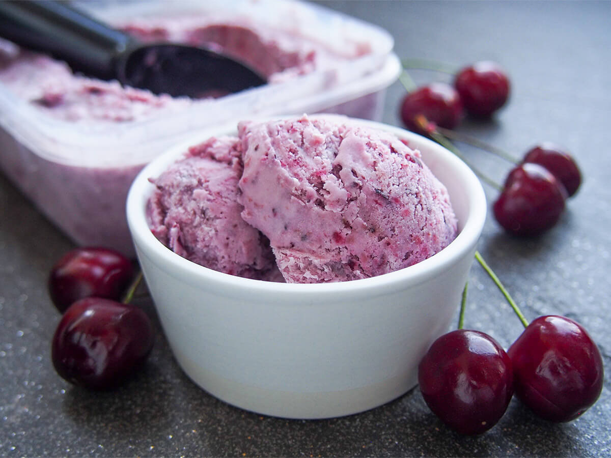 This cherry ice cream is easy to make and packed with wonderfully bright cherry flavor. It uses a simple no churn method that results in a wonderful balance of sweetness, creaminess and fruitiness. bowl of cherry ice cream with cherries around bowl and tub behind