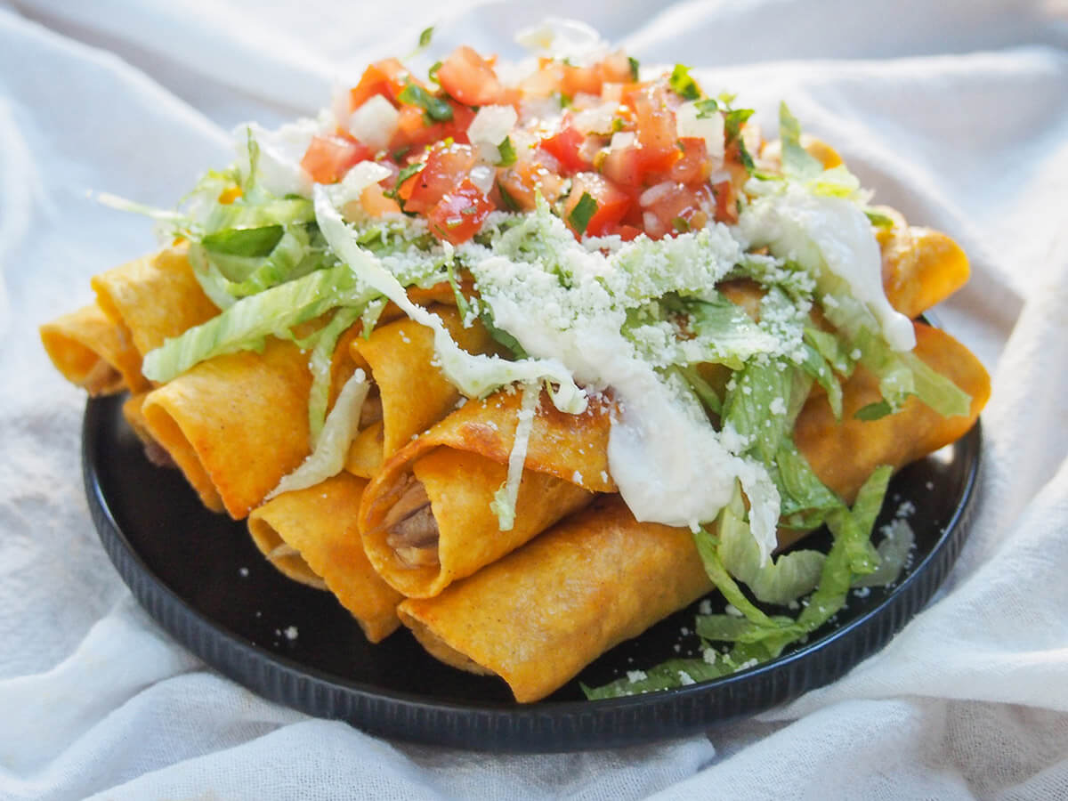 Chicken flautas are a simple combination of tortillas filled with chicken, then fried to crisp deliciousness. Load up with toppings for a great appetizer or snack. plate of chicken flautas loaded with lettuce, pico de gallo, cream and cheese on top