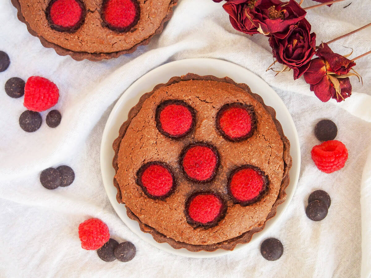 This individual chocolate raspberry almond tart is pretty, elegant and thoroughly delicious. A wonderful sweet treat with great flavors. a chocolate almond tart with raspberry with raspberries and chocolate chips around plate