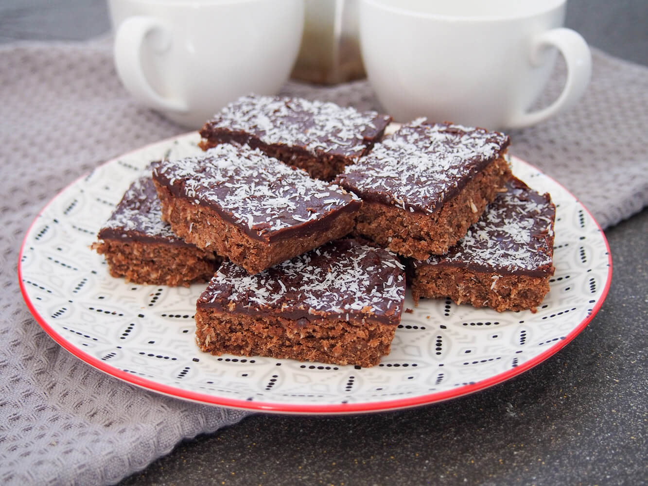 Chocolate slice is an easy to make Australian treat, kind of like a cookie bar with coconut mixed in and topped with a chocolate glaze. It's easy to scale up or down and perfect as an after school or tea time snack. plate of chocolate slice pieces stacked on it