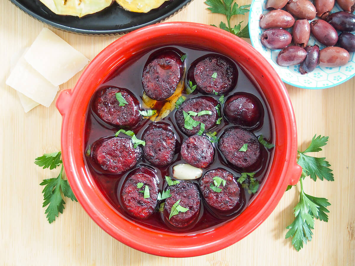 Chorizo al vino tinto is a simple Spanish tapas dish where you cook slices of chorizo in red wine giving them a wonderful extra boost of flavor. They're great to nibble on along with other tapas for a light meal or snack. Chorizo al vino tinto (chorizo in red wine) with olives to one side and cheese to other