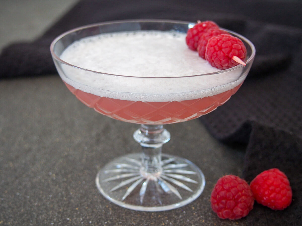 This prettily colored drink is a twist on the classic Clover Club gin cocktail. It has a lovely gently aromatic flavor and is relatively tart, as well as packing a bit of a punch, too. glass of Clover club in Paris - a gin cocktail - topped with raspberries on a cocktail stick