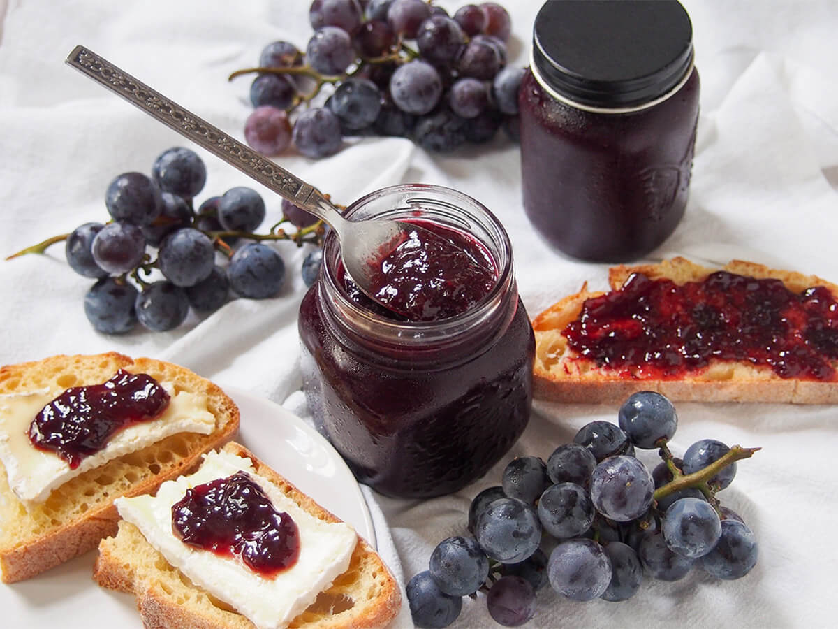 Concord grape jam jar of Concord grape jam with piece of bread topped with jam to one side, and bread with cheese and jam on other