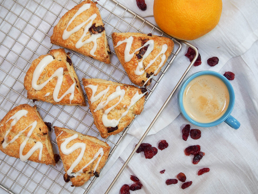 These cranberry orange scones are an easy, tasty treat. With delicate flavors and a tender crumb, they're the perfect snack alongside a cup of coffee or tea. Cranberry orange scones with cup of coffee and orange to side from overhead