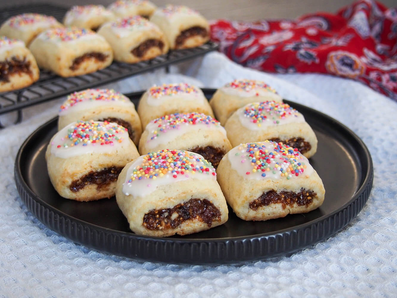 These cucidati have a delicious fruit filling wrapped in a buttery cookie with a flourish of lemon frosting and sprinkles on top. It's no wonder these Italian fig cookies are a favorite. cucidati Italian fig cookies on plate with more on rack behind