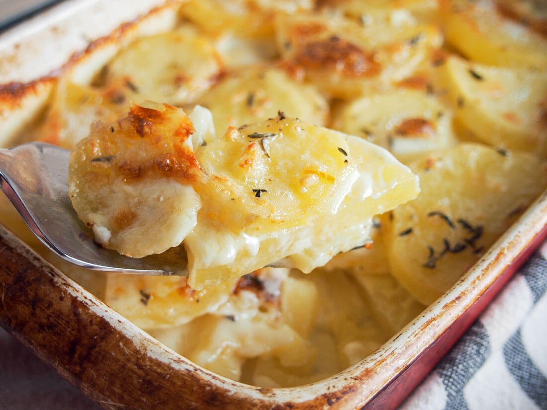 Dauphinoise potatoes are a wonderful combination of creamy potatoes with a touch of garlic and thyme. Rich, meltingly tender and so good. spoonful of Dauphinoise potatoes held in front of dish filled with rest