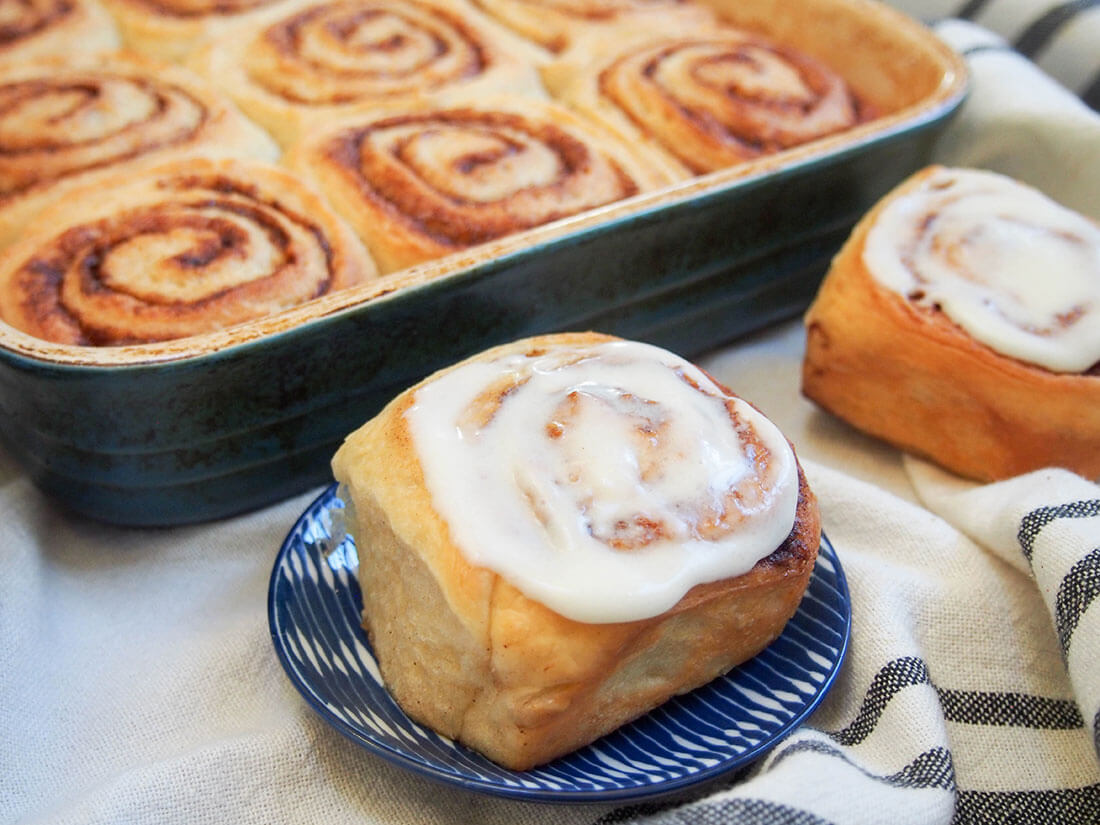Warmly spiced, pillowy soft bites of tastiness, these eggnog cinnamon rolls are just begging to be eaten. They're the perfect addition to any brunch or celebration. frosted eggnog cinnamon roll on plate in front if dish with more