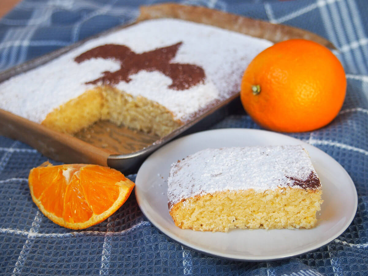 Schiacciata alla fiorentina is a traditional Italian orange cake or sweet bread. It has a lovely flavor and texture and is perfect as a coffee time treat. Traditionally enjoyed during Carnevale (Mardi Gras), it's delicious any time. slice of Florentine orange cake schiacciata on plate in front of rest and orange behind plate