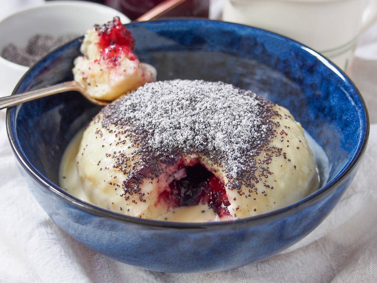 Germknödel are a traditional German/Austrian yeast dumpling, filled with plum butter or jam and topped with poppy seeds and sugar. Germknödel in bowl with spoonful taken from it sitting at back and showing jam in middle.