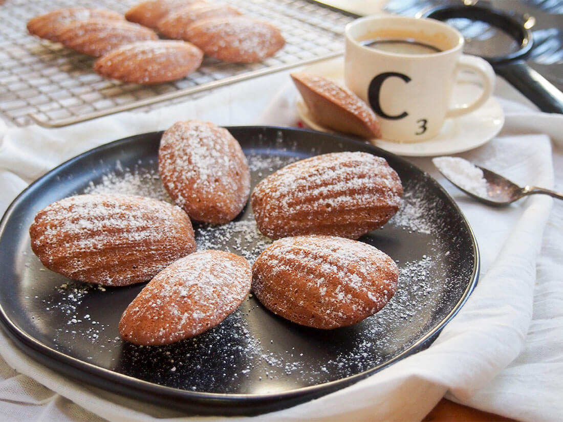 These madeleines are a delicious festive twist on the traditional French cookie. Gently spiced and wonderfully light with a crisp outside, they're little bites of heaven. gingerbread madeleines on plate with coffee cup behind