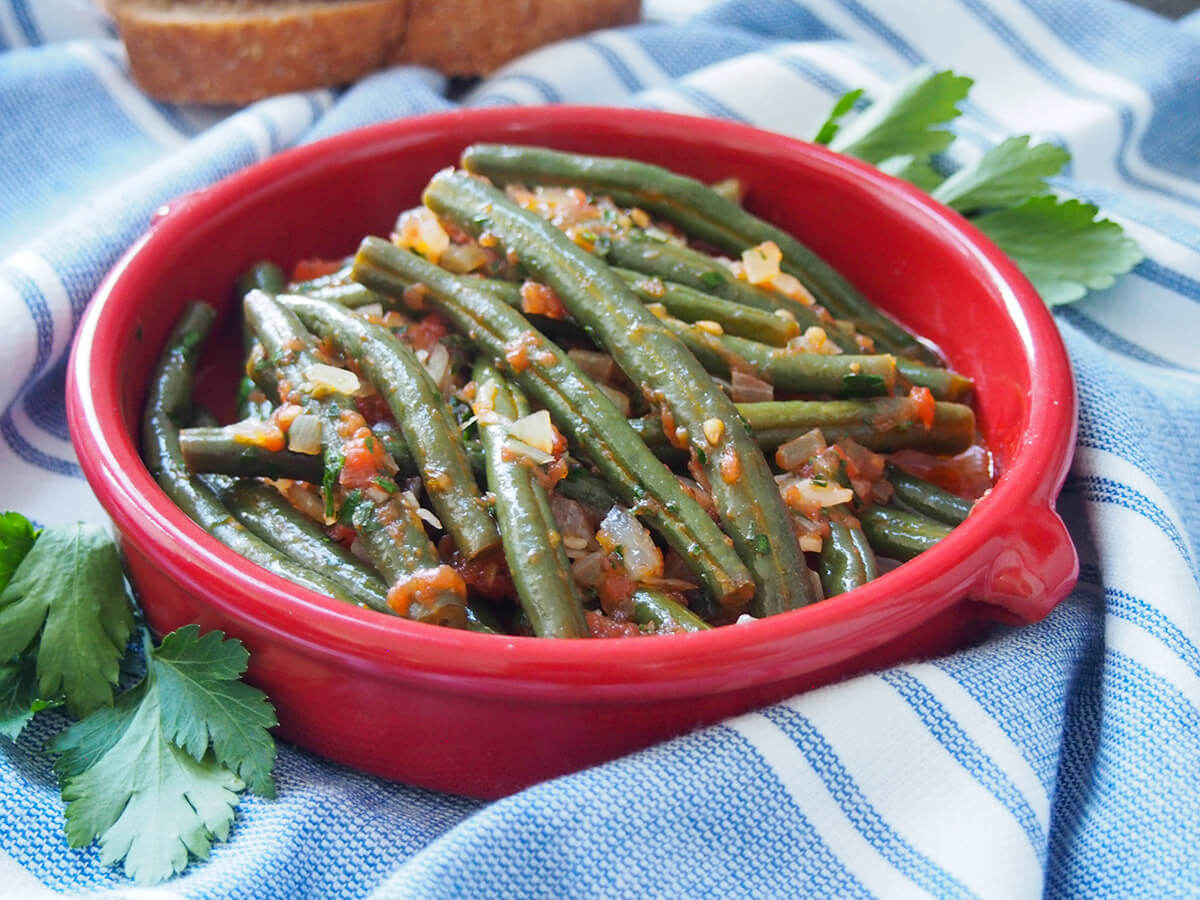 These Greek green beans, fasolakia, are a simple way to cook them, using olive oil and tomatoes, but the result is so fresh, flavorful and delicious. bowl of Greek green beans fasolakia on blue cloth