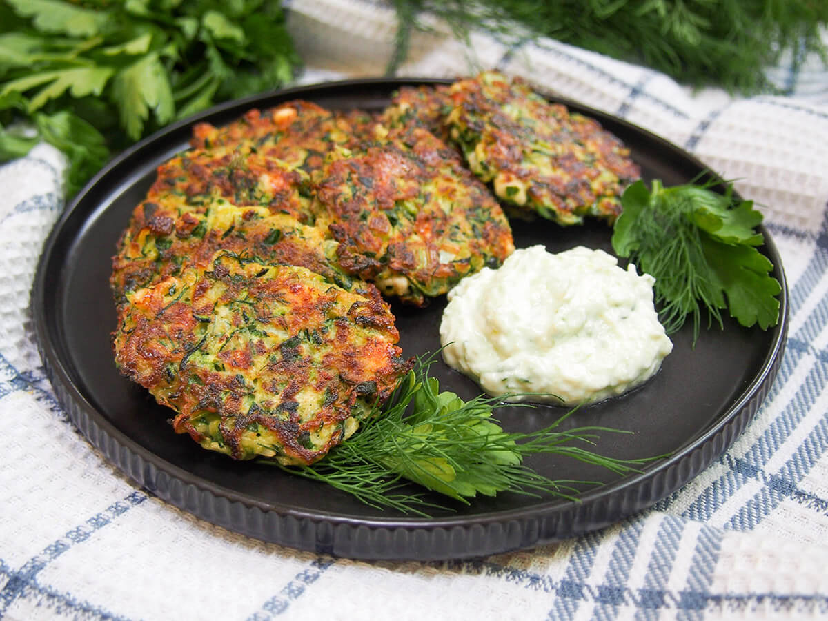 These Greek zucchini fritters, kolokithokeftedes, are a tasty combination of zucchini combined with bright herbs and gently salty feta. They're perfect to snack on or serve as a side, as well as being a great way to use up some zucchini. plate of Greek zucchini fritters (kolokithokeftedes) with herbs at front and yogurt sauce to side