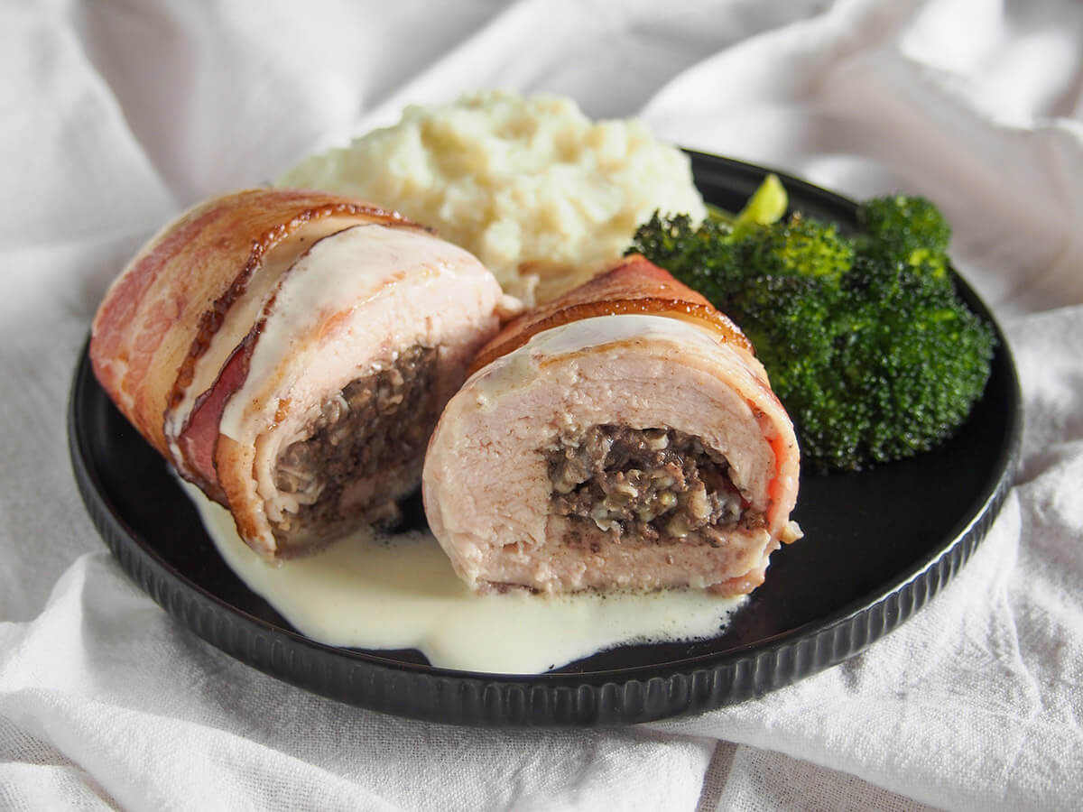 Chicken Balmoral is a Scottish dish of haggis stuffed chicken, wrapped in bacon and served with whisky sauce. It's a delicious combination. black plate with haggis stuffed chicken Balmoral with mashed potato and broccoli behind