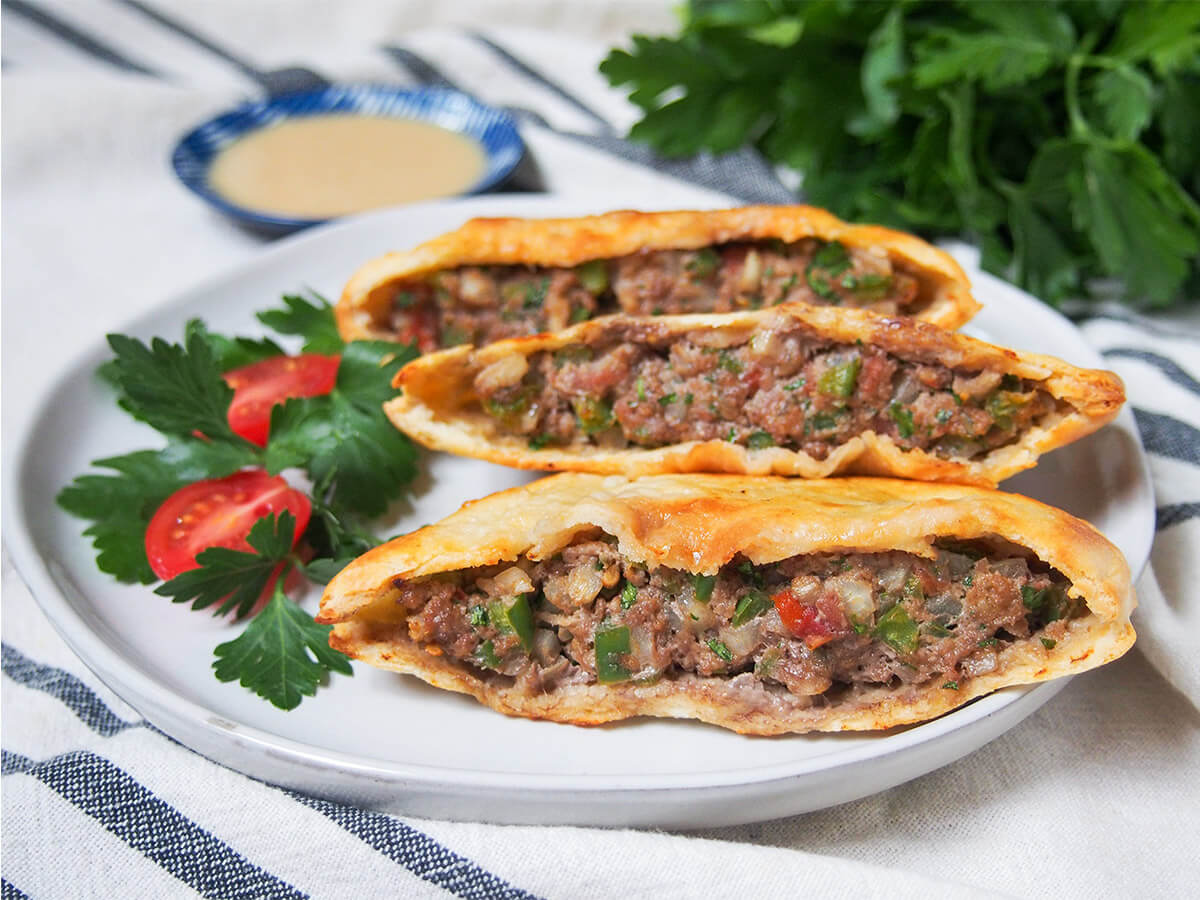 Hawawshi is a traditional Egyptian street food that's essentially a baked ground/minced meat pita sandwich. The spiced and vegetable-loaded meat filling is flavorful and the bread crisps up as it cooks. three hawawshi meat filled pita on plate with tomato and parsley to side