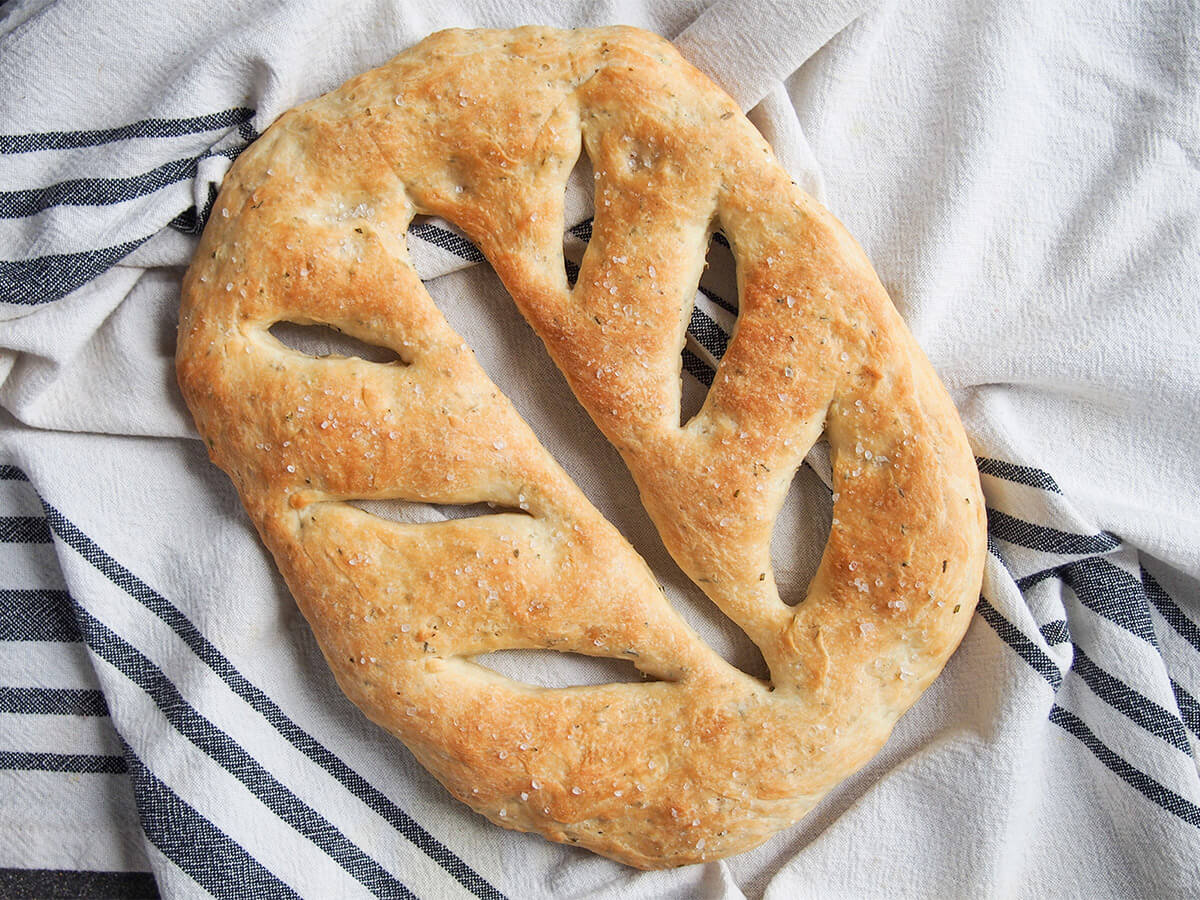 Fougasse is the French cousin of focaccia and you can see the French flair in this pretty and flavorful herb loaf. This herb fougasse comes together more easily than you might think and the result is a delicious aromatic bread. whole herb fougasse on towel