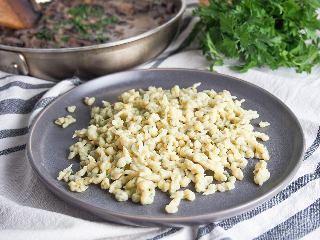 Herb spaetzle are easy to make and tasty both as a side or on their own with butter and herbs & optionally cheese. Comfort food, for sure! herb spaetzle on plate with herbs and dish with mushroom stroganoff behind