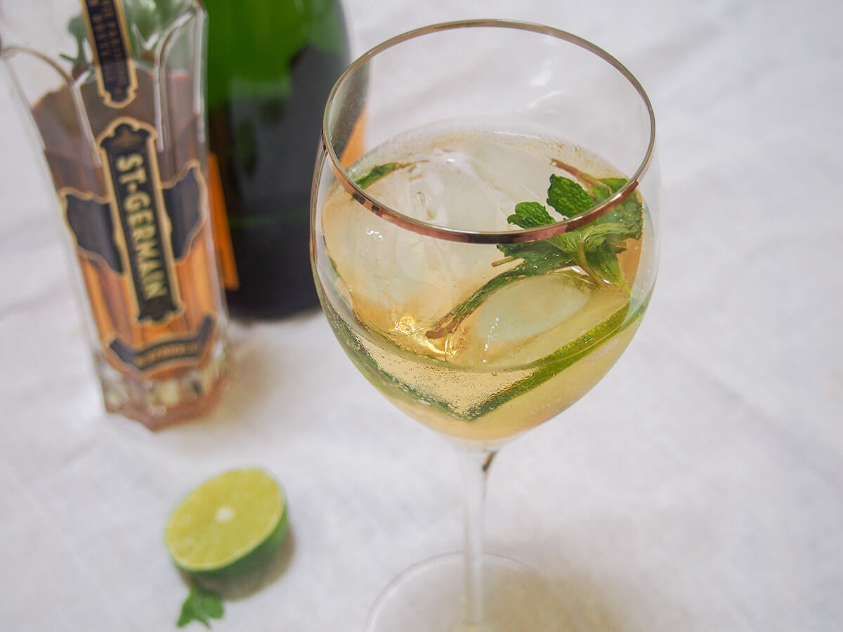 This Hugo spritz cocktail is easy to make, with a bright, refreshing flavor. It has a touch of aromatic sweetness from the St Germain, gentle bubbles and is nicely light. Perfect summer sipping. glass of Hugo spritz cocktail with bottles of prosecco and st Germain behind