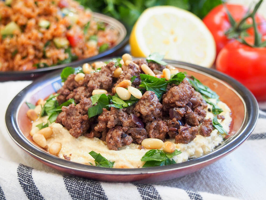 Whether you make your own or buy from the store, it's easy to make your hummus more of a meal with this easy hummus with lamb recipe. The warm spices make the topping so tasty! Hummus kawarma (hummus with lamb) in small dish with tomatoes and plate of kisir behind