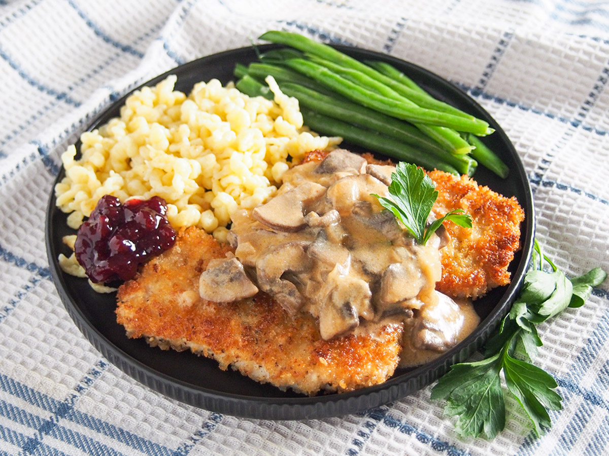 Jaegerschnitzel is the German classic 'hunter's cutlet' AKA a pork chop with a flavorful mushroom sauce. It's comforting deliciousness. plate of jaegerschnitzel with spaetzle and beans behind