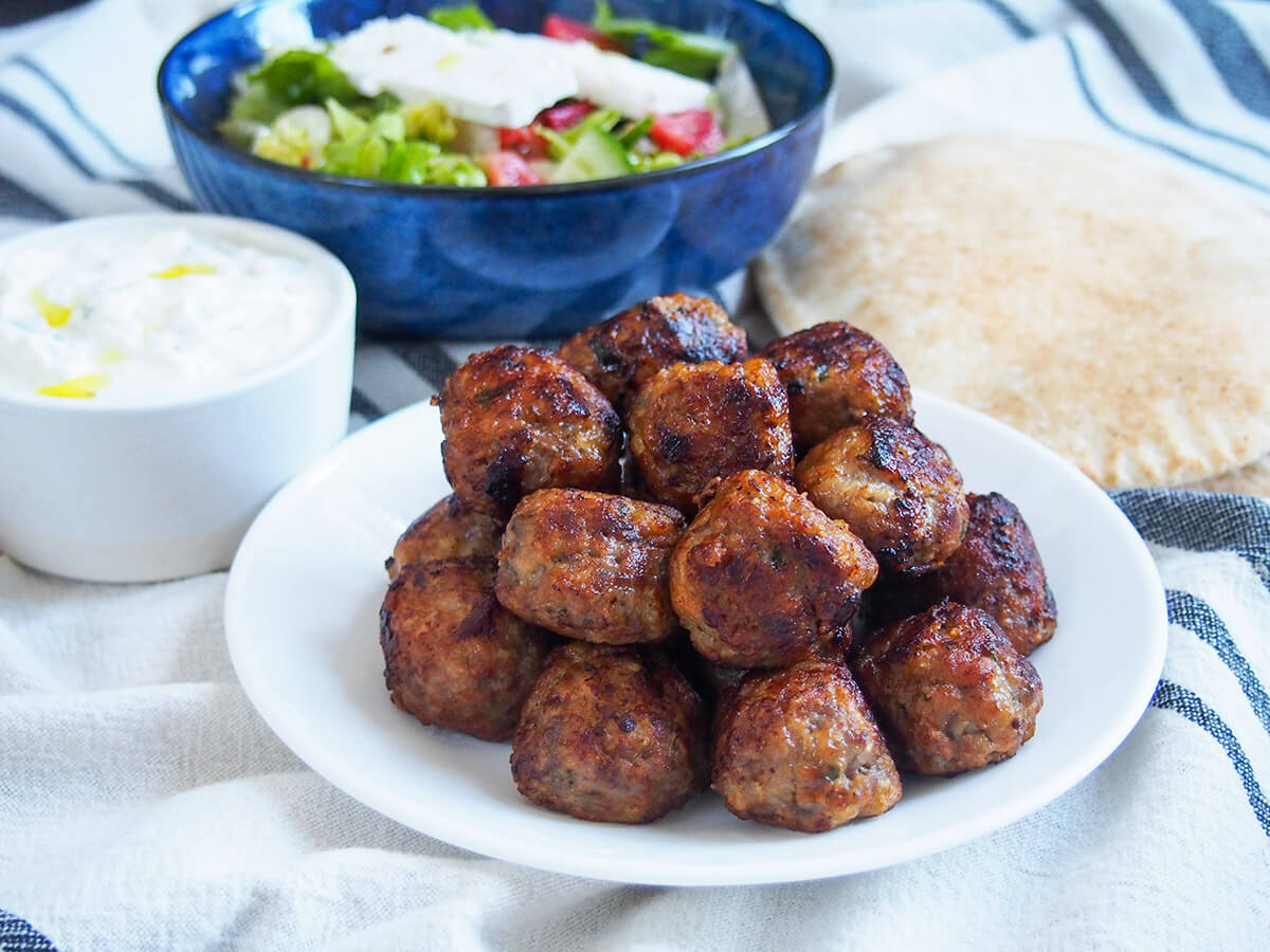 Keftedes are Greek meatballs that are packed with delicious flavor. They are juicy and aromatic with mint and perfect to snack on with other mezze, or enjoy with tzatziki and bread as a wrap. plate of Greek meatballs with tzatziki, salad and pita bread behind