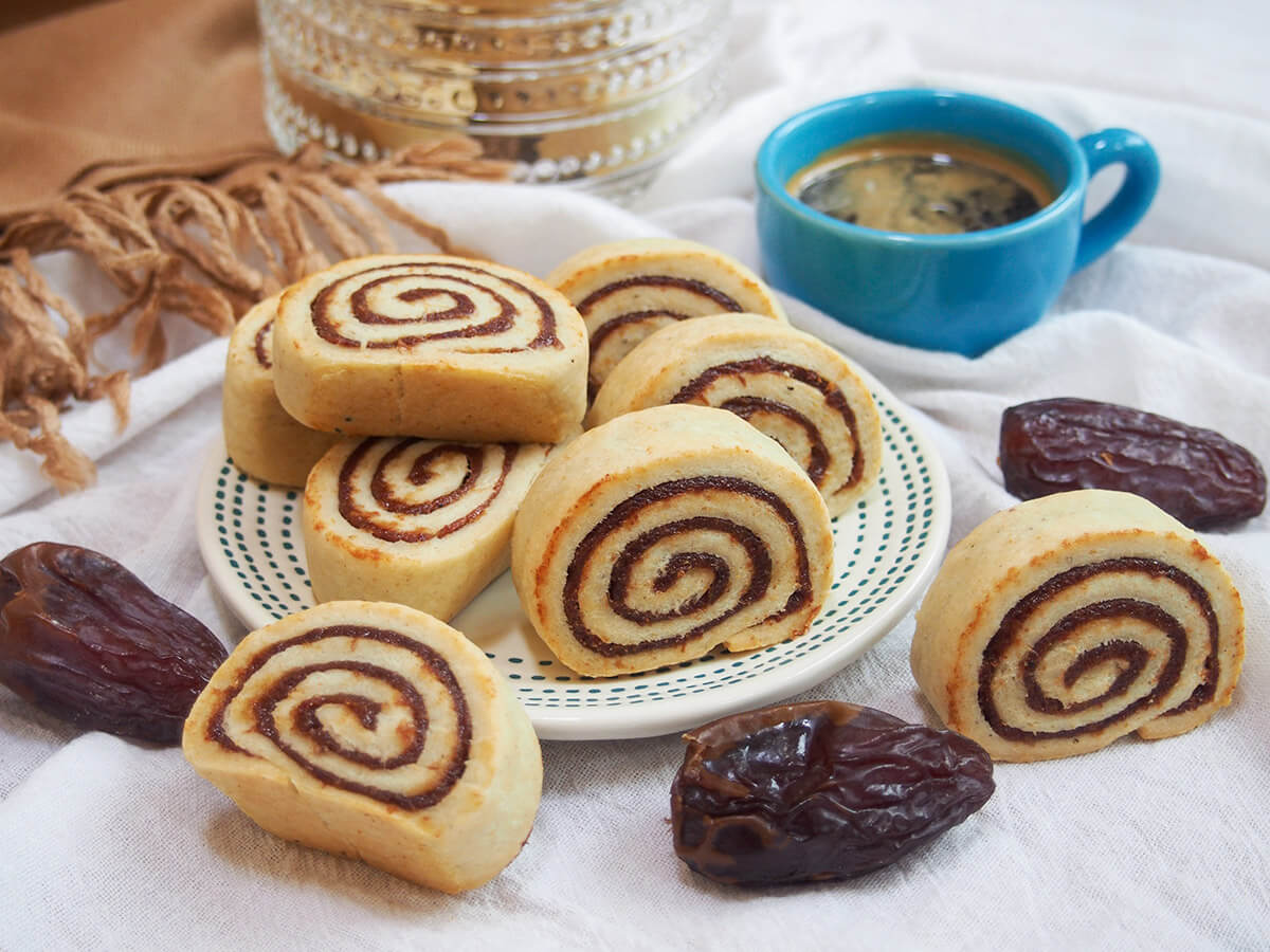 you haven't heard of kleicha, these Iraqi date cookies are a bit like fig Newtons but with a wonderfully aromatic cardamom undertone. And prettier, too, with their elegant spiral shape. They're addictively good! plate of kleicha Iraqi date cookies with coffee cup behind and dates around plate