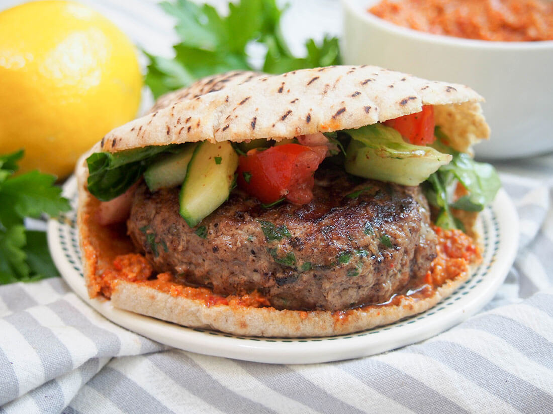 This lamb kofte burger is the perfect burger for when you want something that bit different. Not only are the flavors delicious, but you can cheat a little with ready-made components, or make it more from scratch as time allows. lamb kofte burger with muhammara and lemon in background