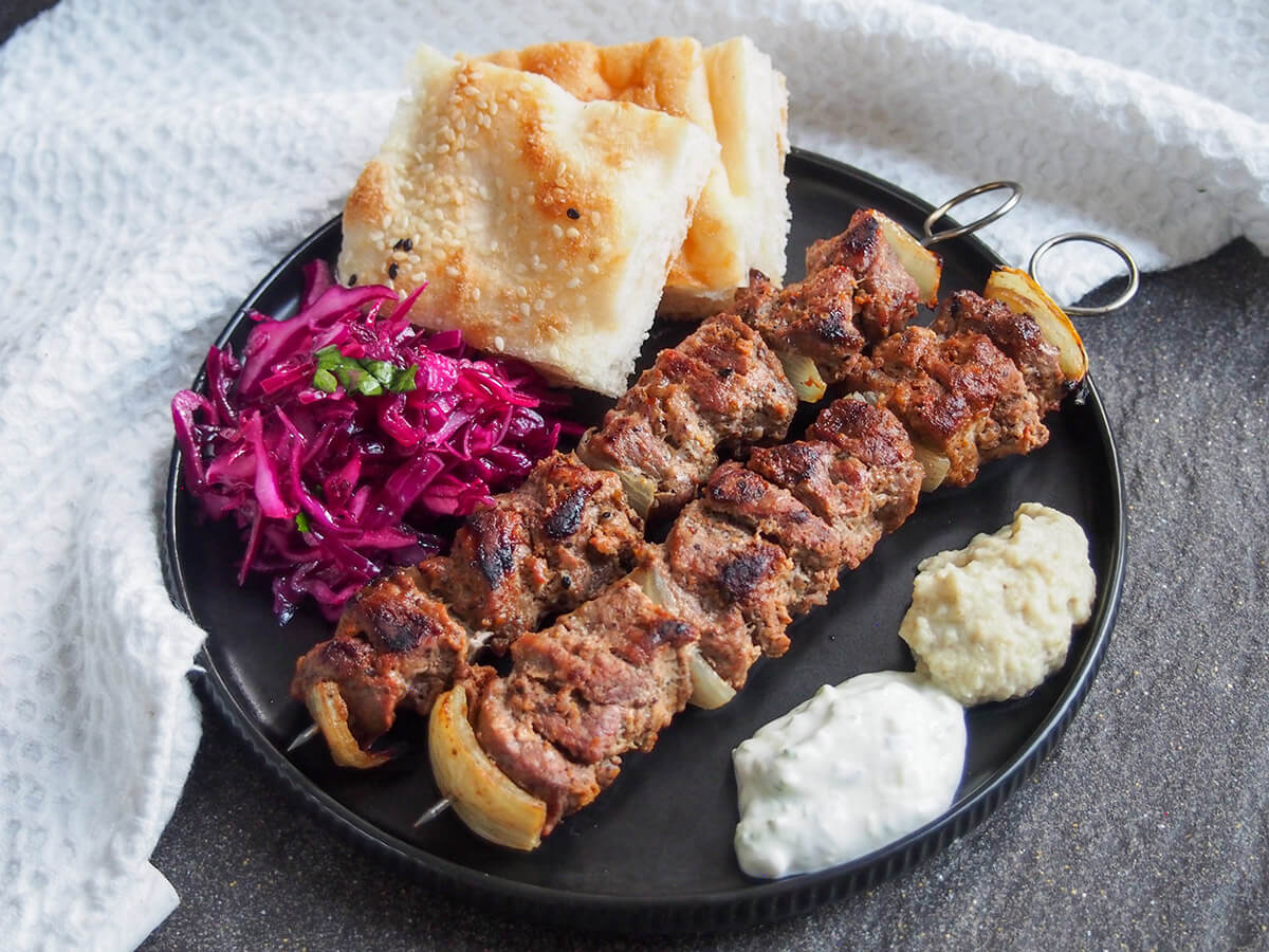 This lamb shish kebab is easy to prepare, deliciously tender from the yogurt marinade with a tasty, light spicing. Pair with various sides for a tasty meal. plate with lamb shish kebab, red cabbage, bread and sauces to side
