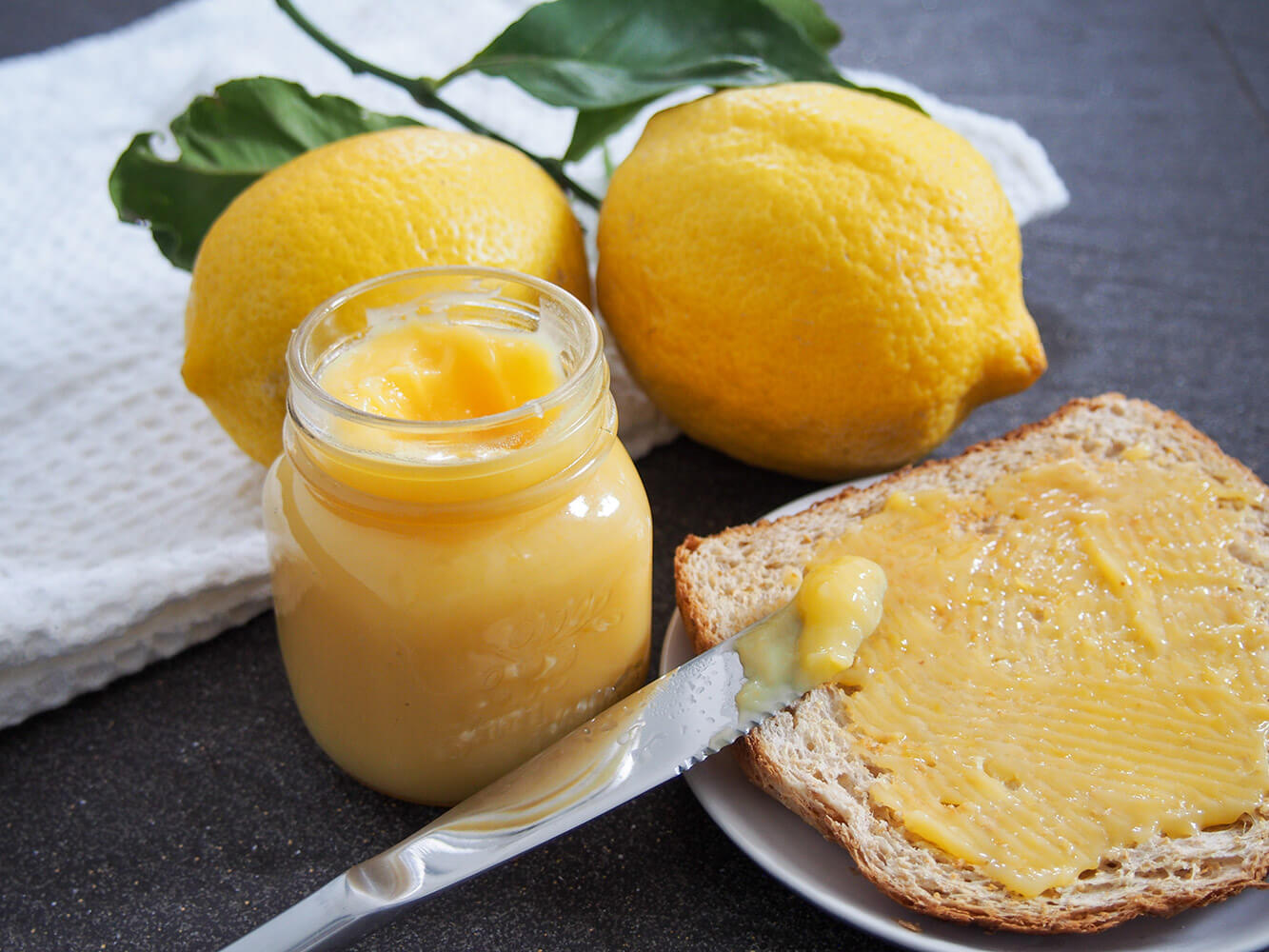 Lemon curd is a delicious spread to put on toast, but also a great ingredient in baking such as for tarts and on pavlova. Slightly sweet with a fantastic lemon zing, it's easy and delicious! jar of lemon curd with some on toast to side and lemons behind